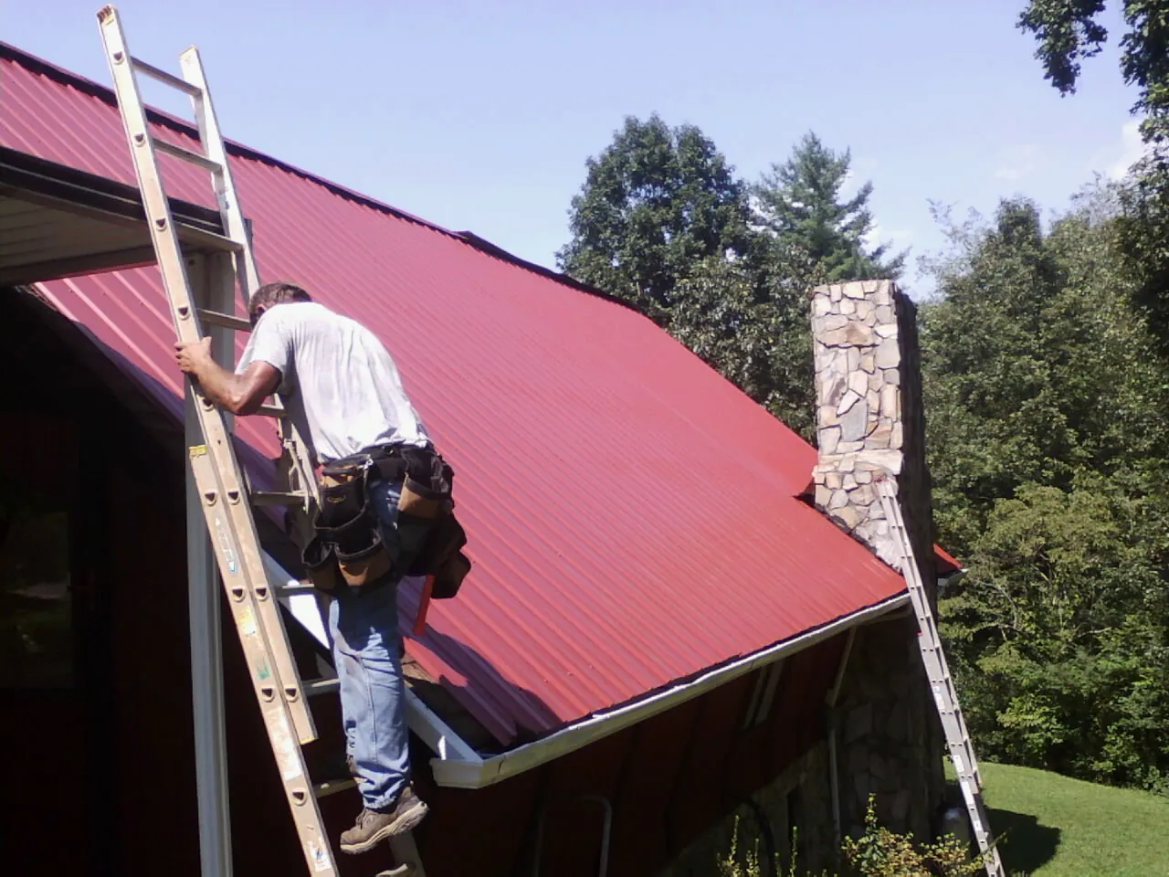 Roofer on a ladder beside a red metal roof and stone chimney — Soffit & Fascia Repair by Roofing Brothers in Dunn Loring